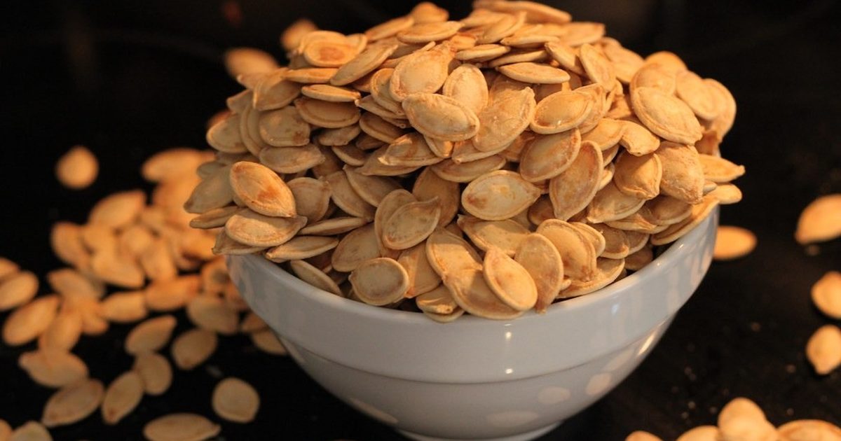 A white bowl heaped with roasted pumpkin seeds on a dark table