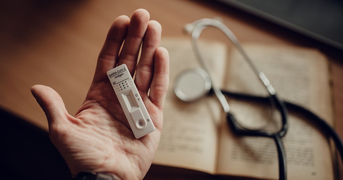 A hand holds a home test device next to a stethoscope and an open medical notebook