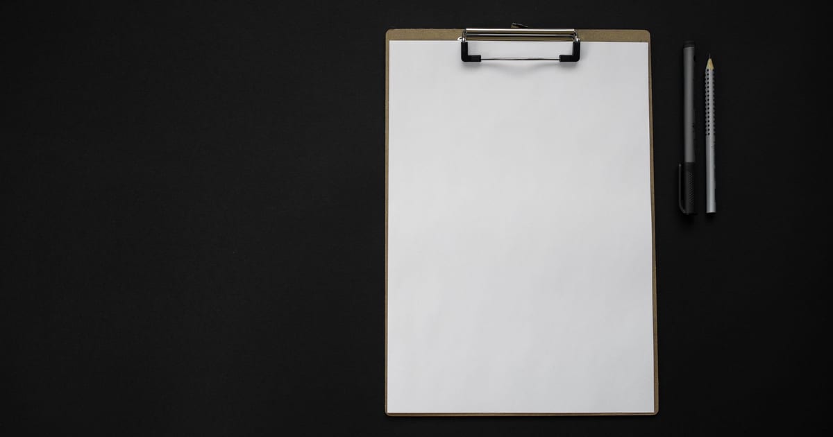 A blank sheet of paper on a clipboard next to a pen and a pencil on a black surface