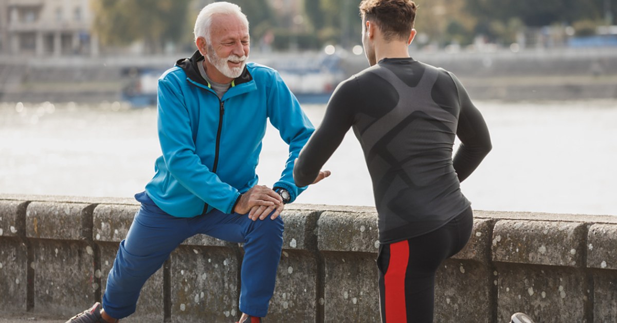 An older man in a blue jacket does a lunge stretch beside a younger man on a riverside walkway