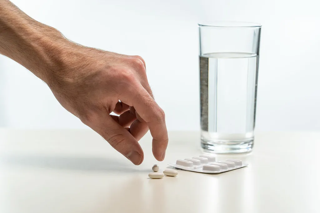 A man's hand reaching for white tablets beside a blister pack and a glass of water