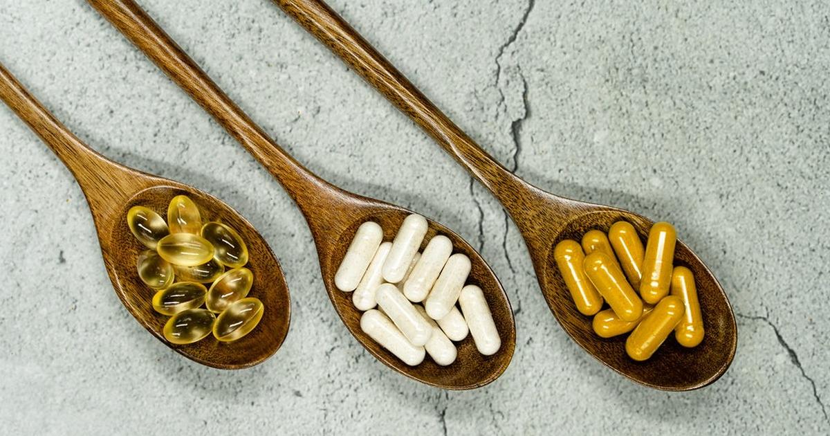 Three wooden spoons holding softgels, white capsules and yellow capsules on a gray stone surface