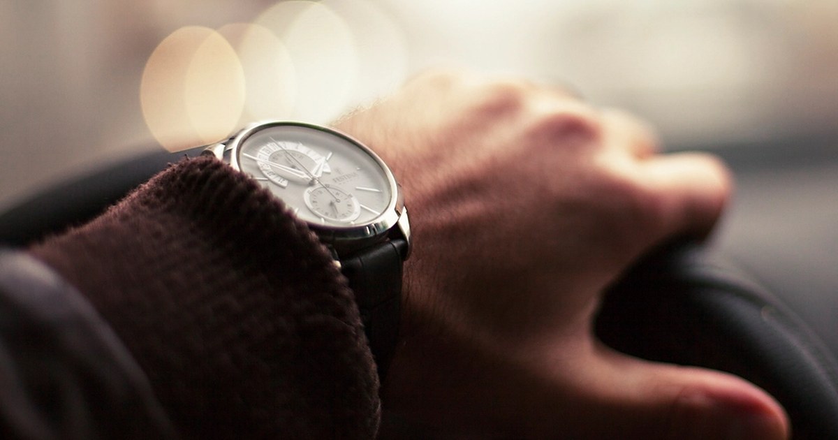 A man's hand on a car steering wheel, with an analog wristwatch on his wrist